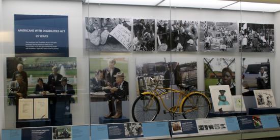 he Americans with Disabilities Act, 1990-2015 artifact wall, showing large graphics of the signing of the law by Pres. Bush, a yellow bike, and other artifacts behind glass on display in a shallow, tall case